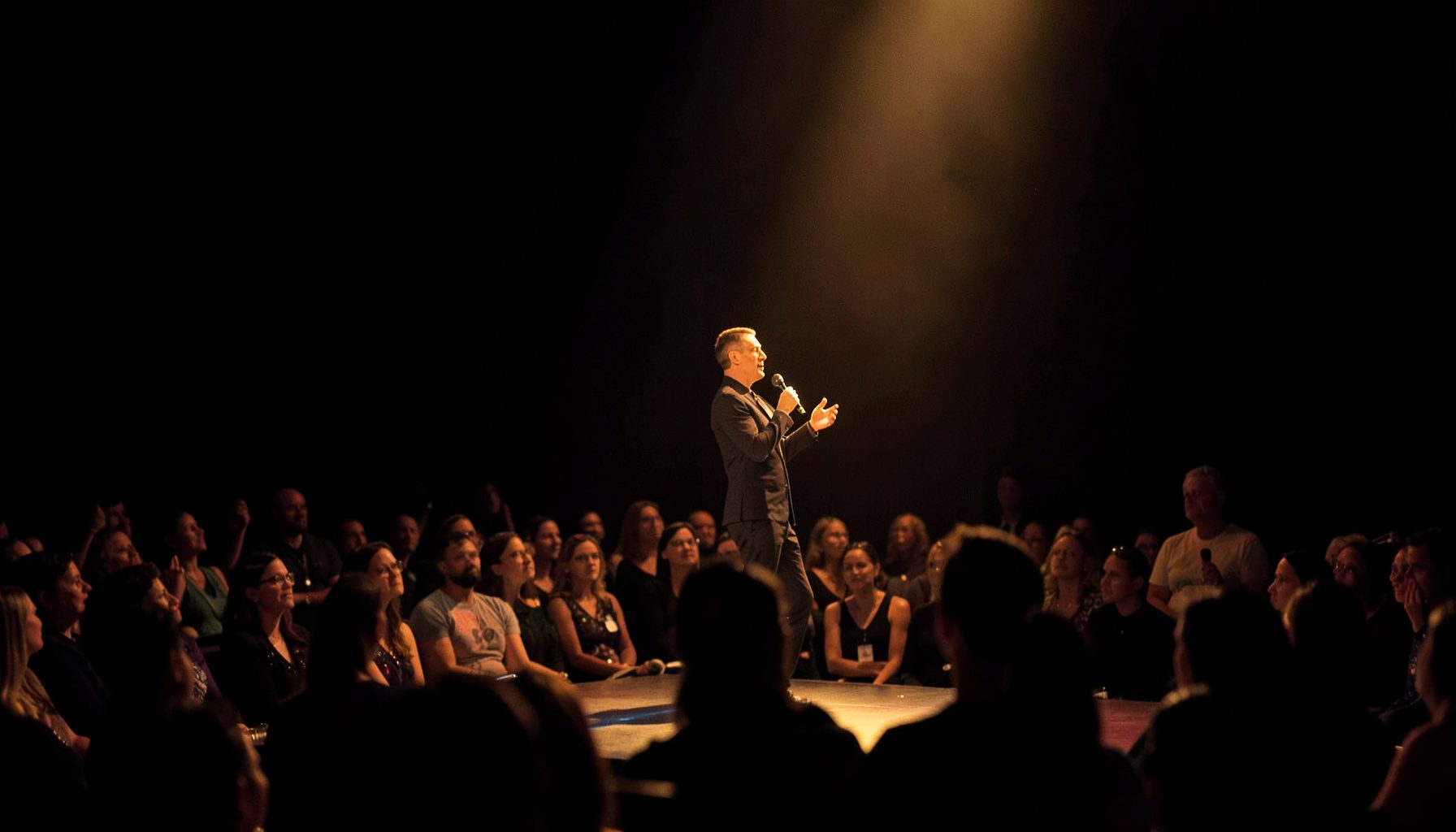 A speaker on stage addressing an audience, warm spotlight on a dark background